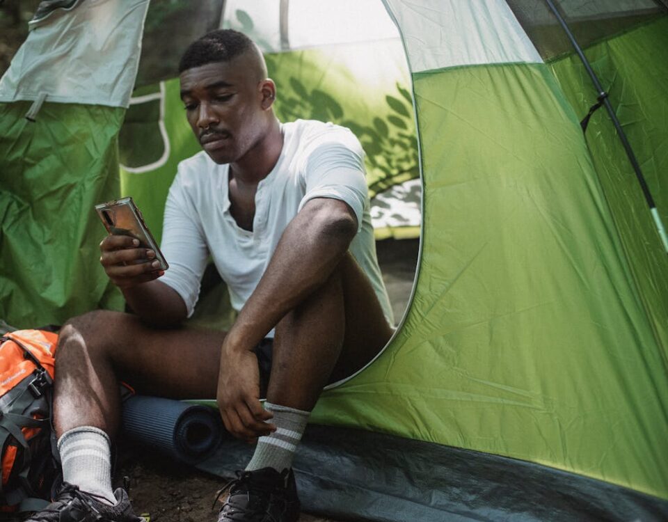 Gadgets essenciais para sua viagem à Bahia Sad African American male traveler resting in tent and browsing Internet on cellphone while spending journey in nature