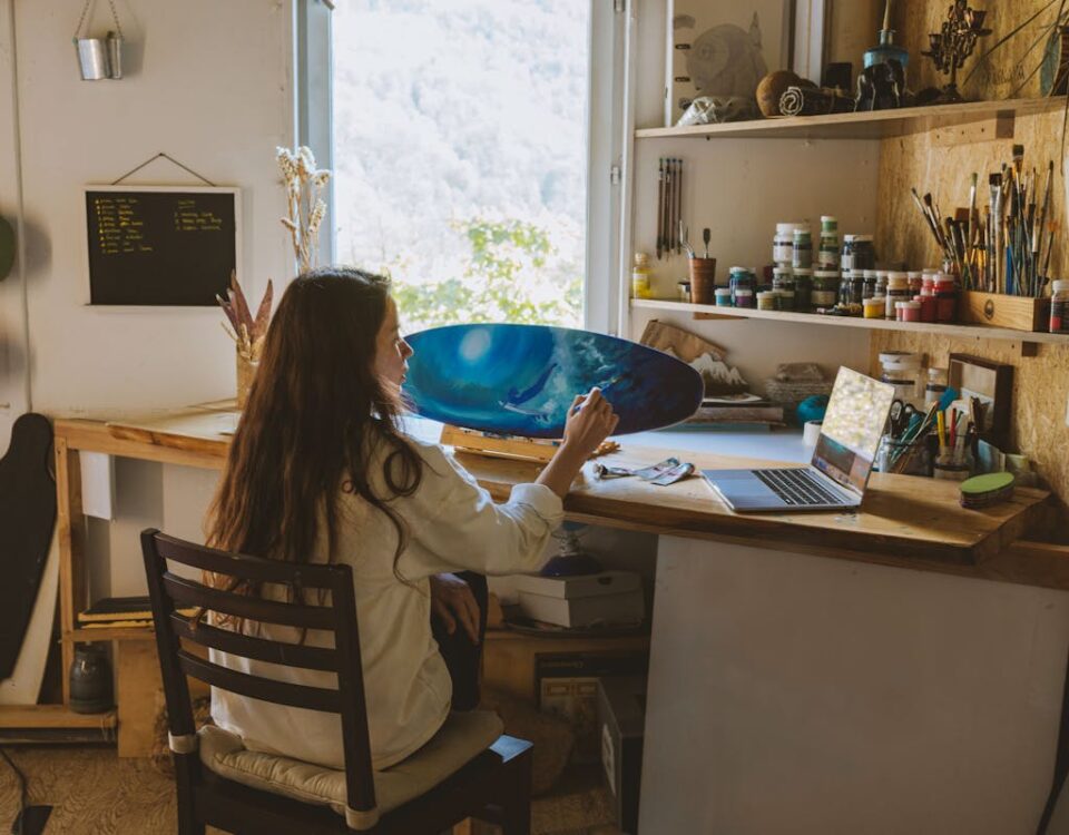 Dispositivos para melhorar a prática de arteterapia Artist painting in a home studio, surrounded by paints and a laptop, showcasing creative workspace.