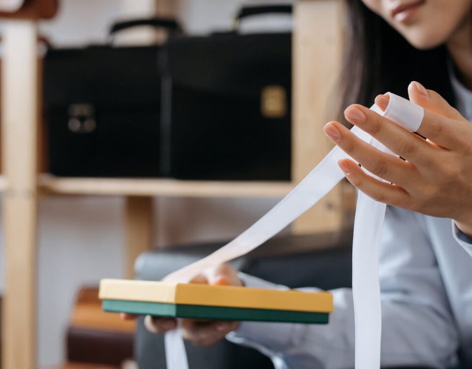 Close-up of a woman holding a gift box with ribbon in an office setting, with briefcases in the background.