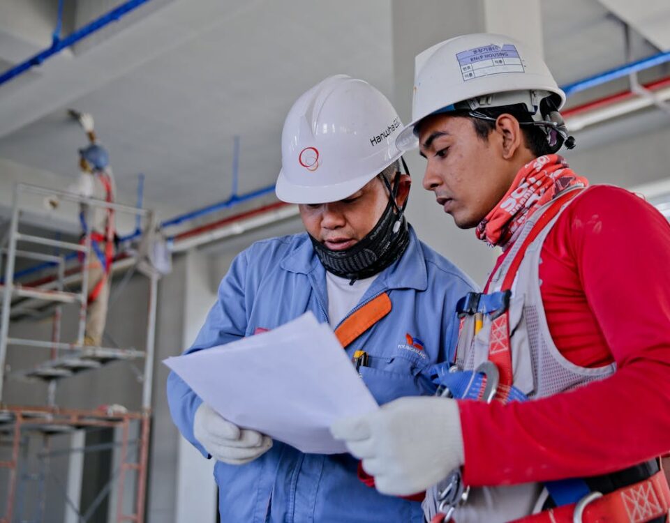 Equipamentos que revolucionaram o cotidiano industrial e doméstico Two engineers in safety helmets reviewing construction plans at a worksite.