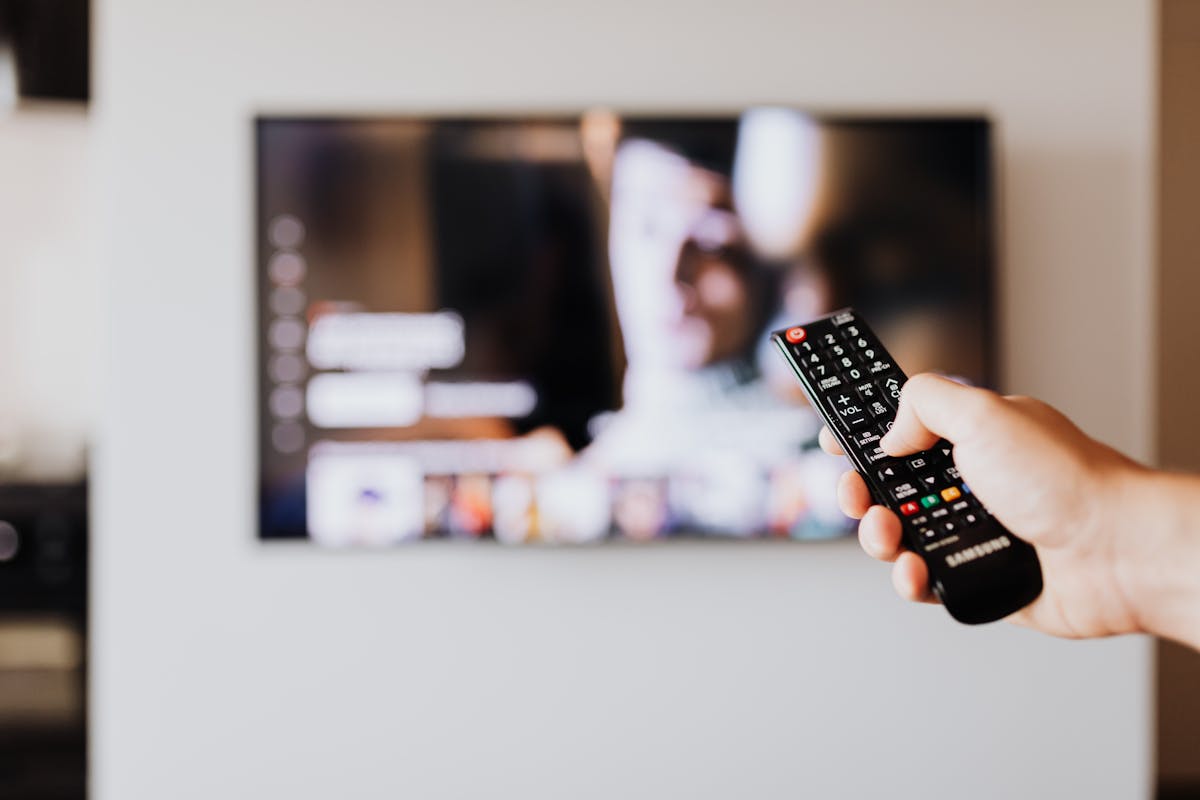 Smart TVs e o futuro do cinema em casa Person using a TV remote control with a blurred television screen in the background.