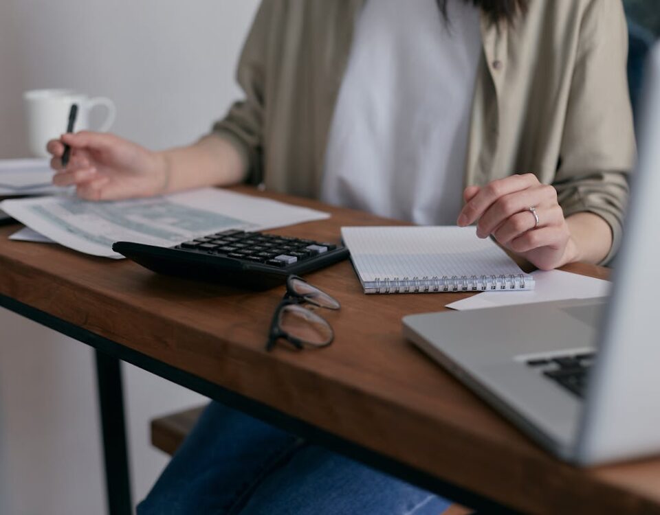 A woman manages finances at home, using a laptop and calculator on a wooden desk.
