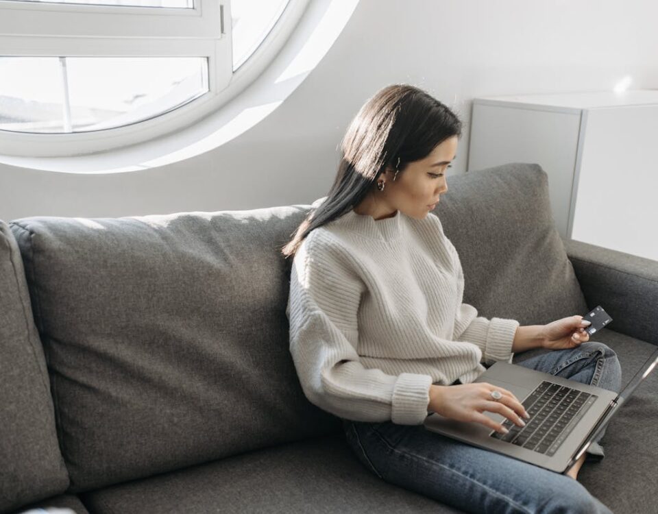 Como comprar eletrodomésticos e acumular milhas? Saiba como Woman sitting on a sofa using a laptop for online shopping indoors, holding a credit card.