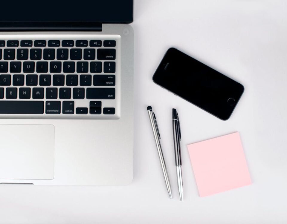 Gadgets que agilizam o dia a dia no escritório A top view of a modern workspace featuring a laptop, smartphone, pens, and a pink sticky note on a white desk.