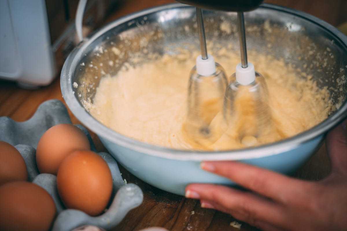Qual a melhor batedeira para massas pesadas? Close-up of batter being mixed in a bowl with eggs nearby on a wooden table.