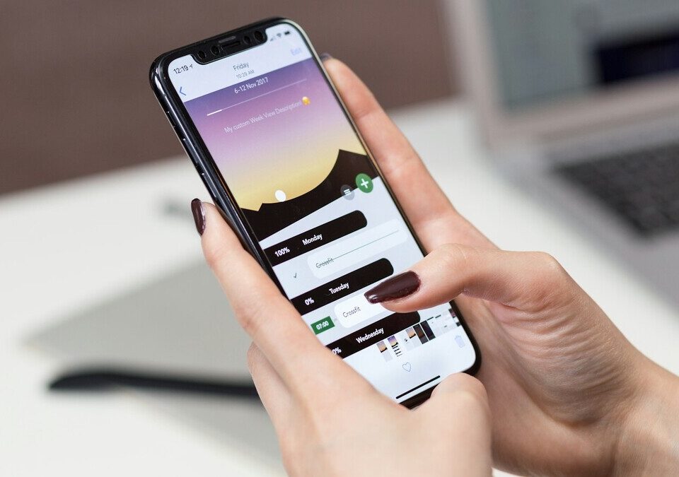 Mão segurando um iPhone com aplicativo aberto, com notebook desfocado ao fundo